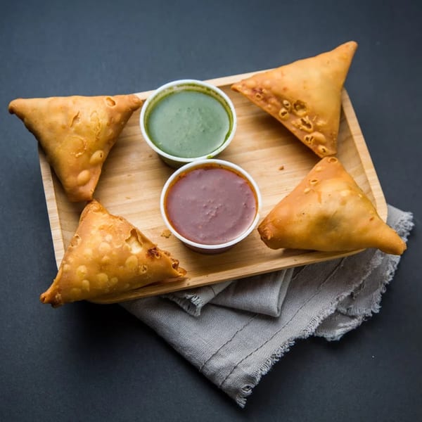 Four golden-brown samosas and two dipping sauces, green and red, on a wooden tray.