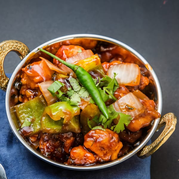 Indian chili chicken with bell peppers, onions, and a green chili in a metal bowl.