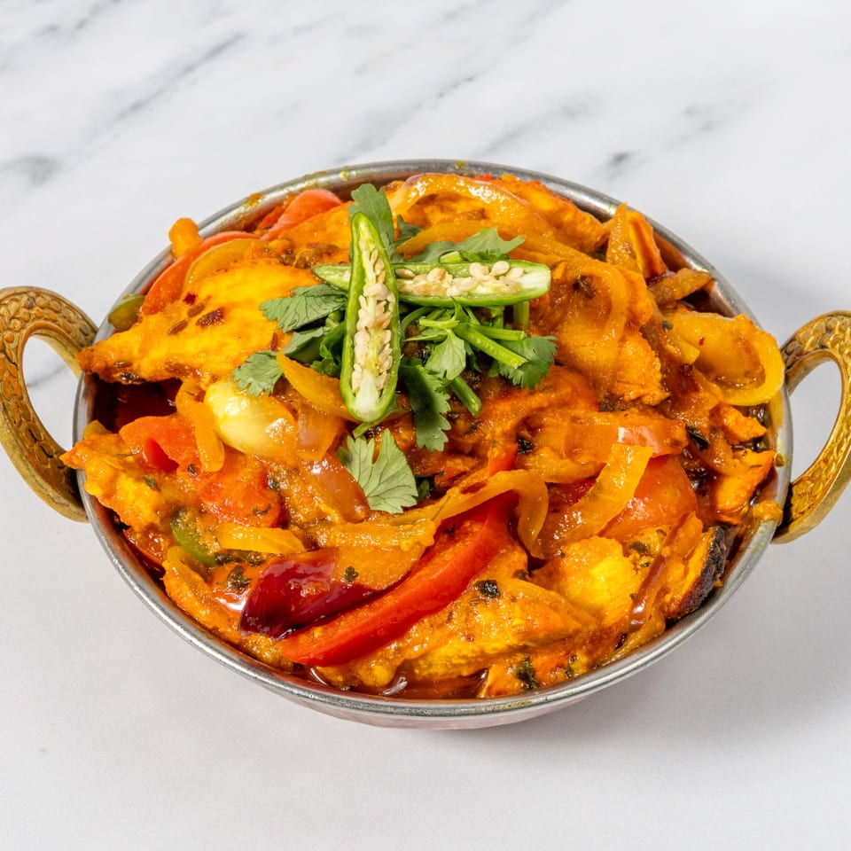 A bowl of richly spiced curry with paneer, tomatoes, peppers, onions, cilantro, and sliced green chilies.