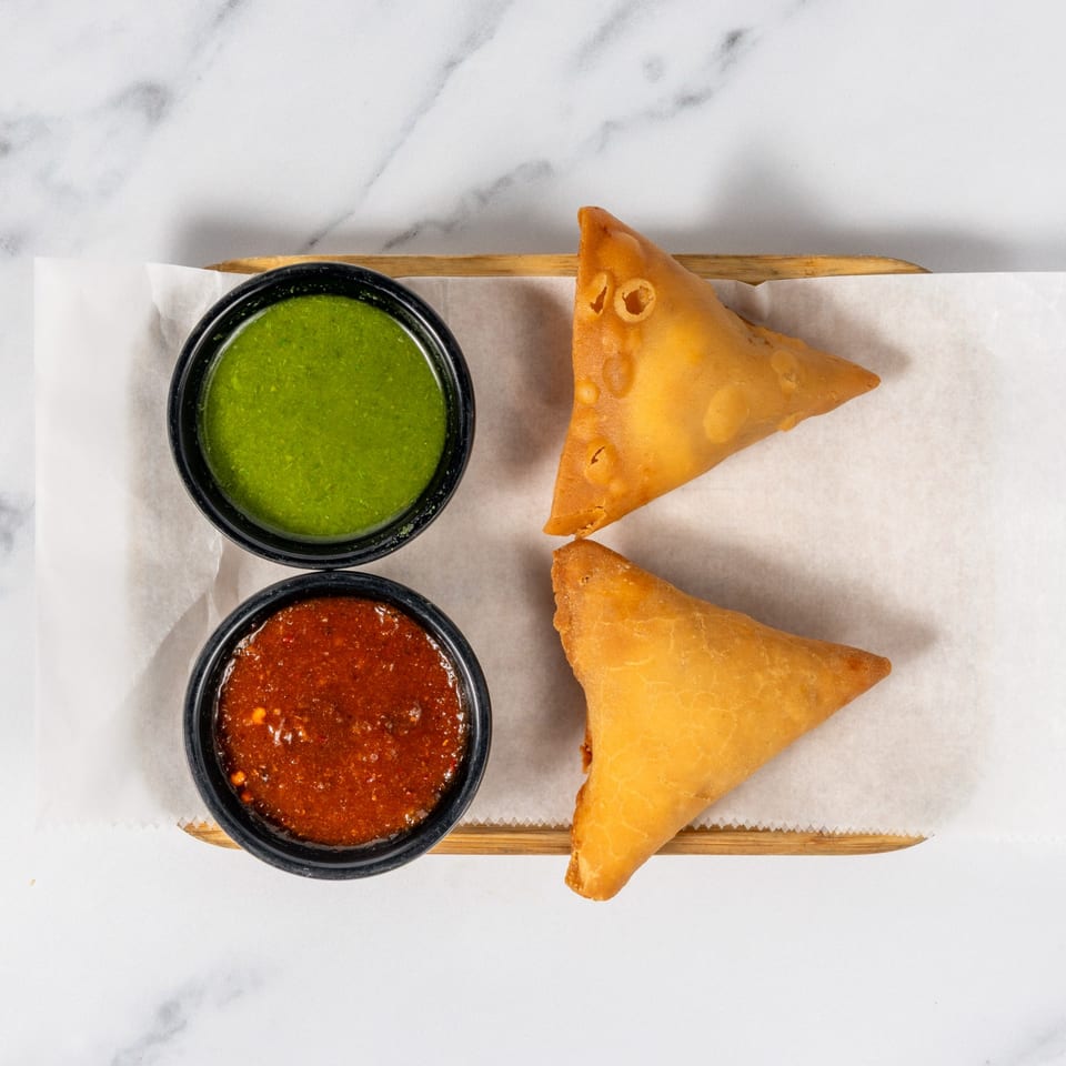 Two golden samosas served with green chutney and red dipping sauce on a paper-lined wooden tray.