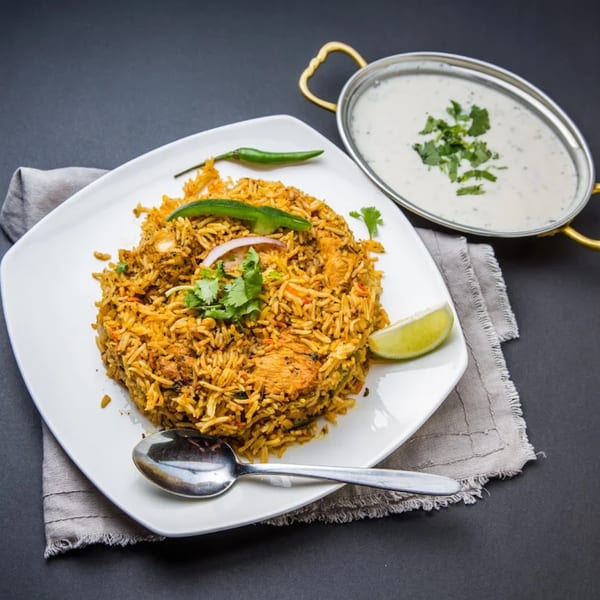 Chicken Biryani with raita, garnished with cilantro, lime, and green chili on a white plate.