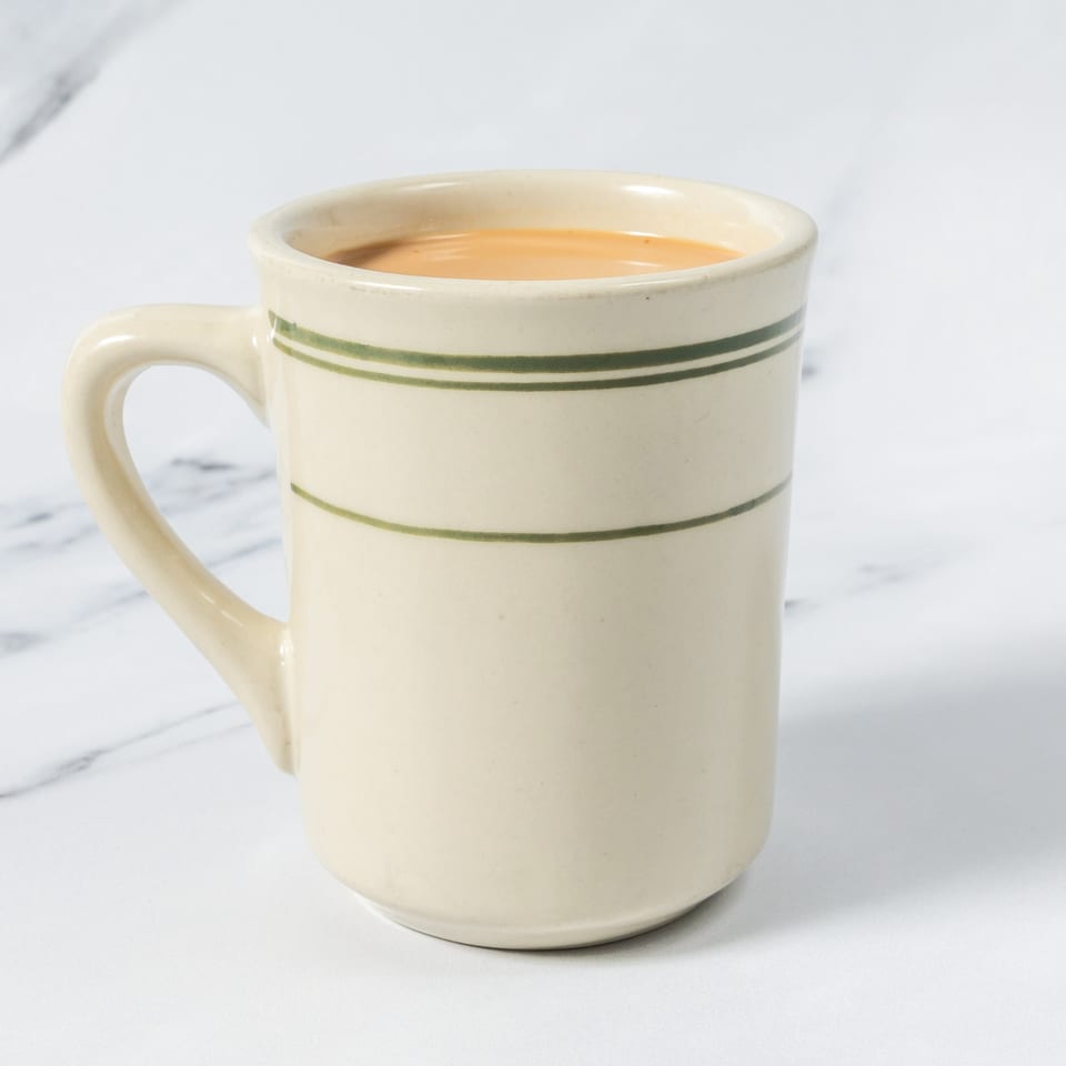 Cream-colored ceramic mug with green stripes, filled with a light brown beverage, sits on a white marble surface.