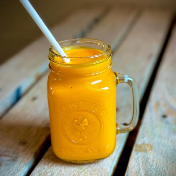 A vibrant orange mango lassi in a mason jar with a white straw on a rustic wooden table.